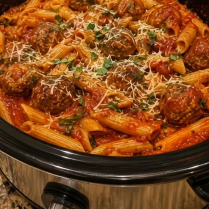 A steaming crockpot filled with penne pasta and meatballs in a rich red marinara sauce, garnished with fresh parsley.