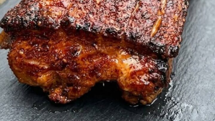 A close-up shot of a perfectly smoked pork belly with a dark, caramelized bark on a slate board.