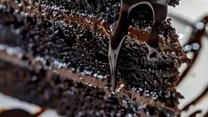 A slice of moist black velvet cake on a white plate with glossy dark chocolate ganache dripping down the sides.
