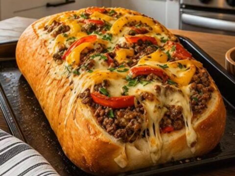 A golden-brown stuffed French bread loaf on a dark baking sheet, overflowing with melted cheese and peppers.
