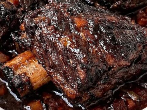 A close-up of tender, glazed beef short ribs with a dark, caramelized crust resting in a white slow cooker.