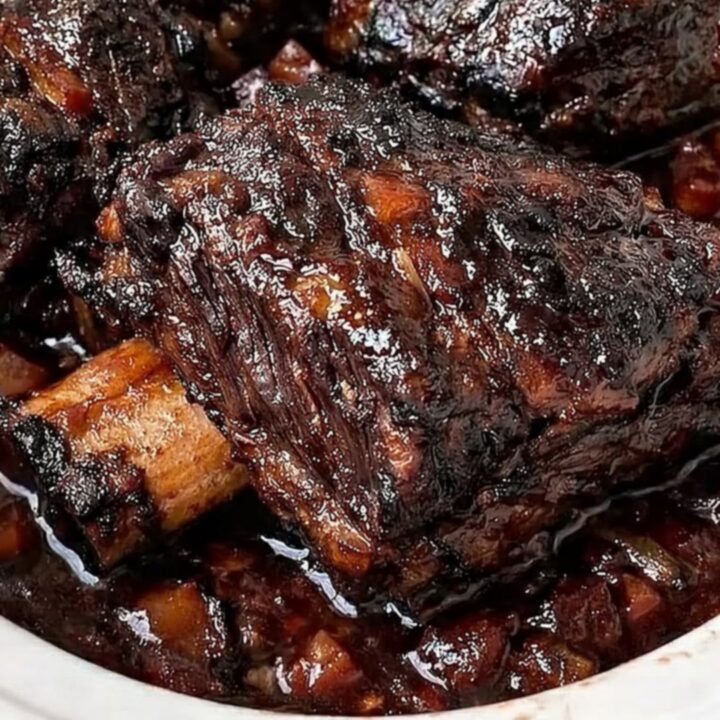 A close-up of tender, glazed beef short ribs with a dark, caramelized crust resting in a white slow cooker.