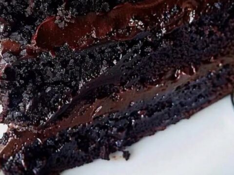 A silver fork cutting into a decadent three-layer dark chocolate fudge cake on a white plate.