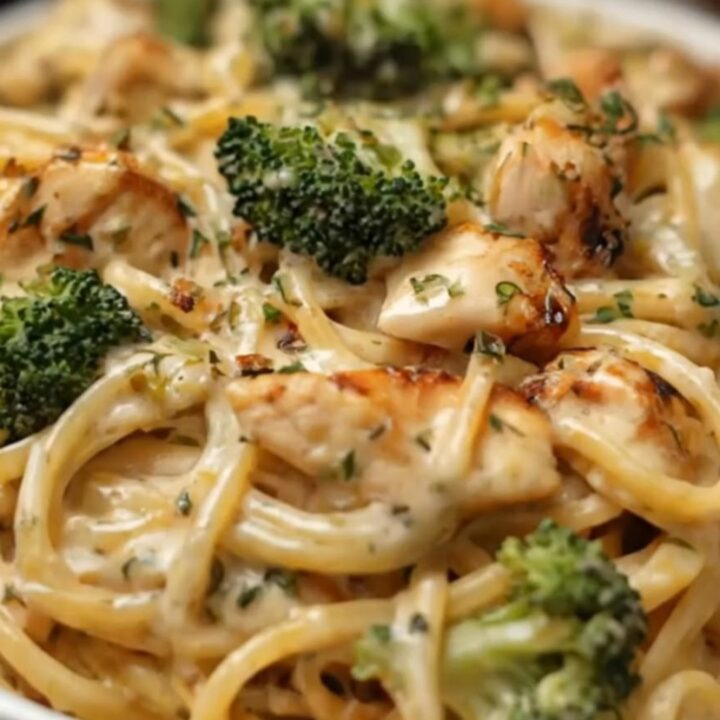 A close-up of a fork lifting creamy linguine, seared chicken pieces, and vibrant green broccoli from a fluted white bowl.