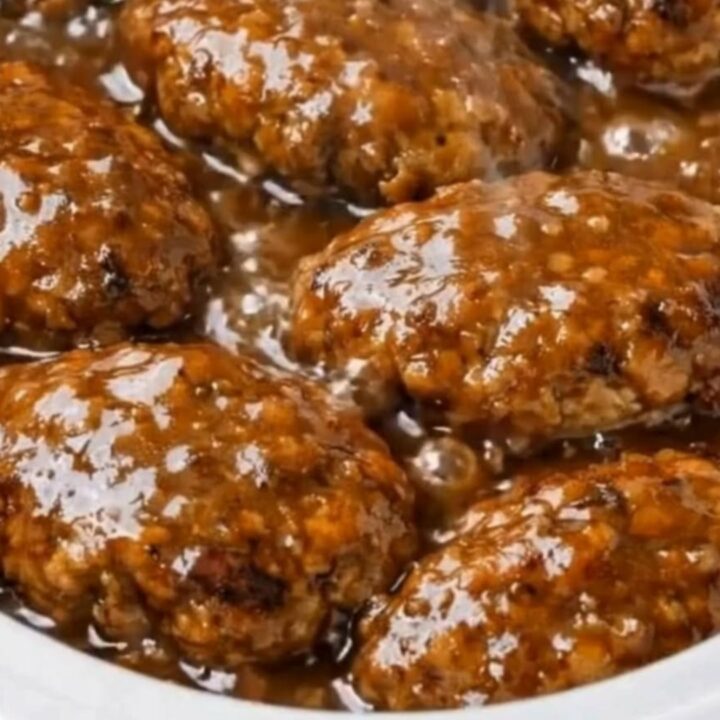 Six glistening Salisbury beef patties gently bubbling in a rich, savory onion and mushroom gravy inside a white slow cooker.
