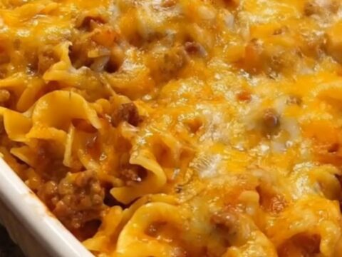 A hearty serving of beef noodle casserole being lifted from a white baking dish with a dramatic cheese pull.