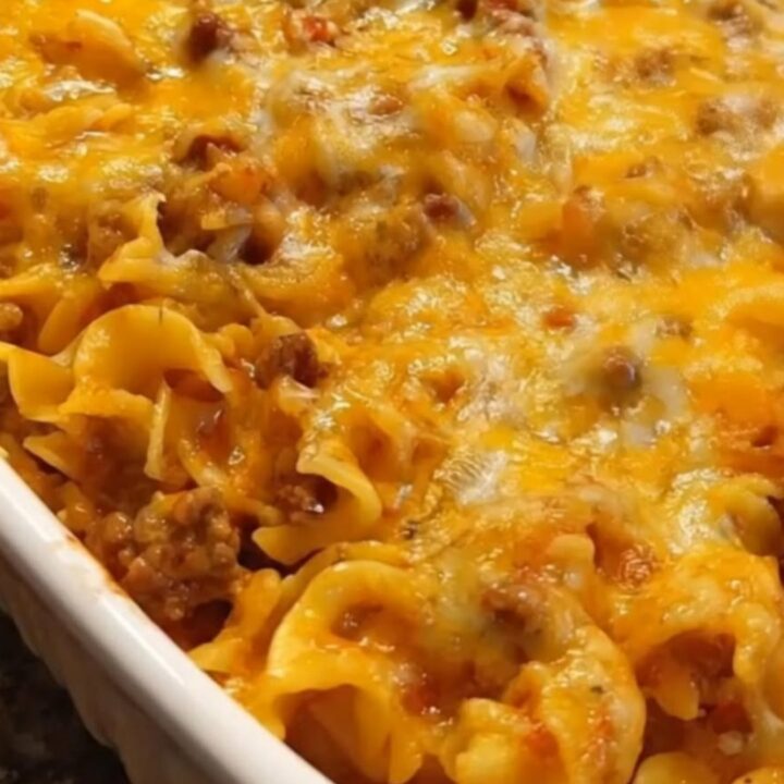 A hearty serving of beef noodle casserole being lifted from a white baking dish with a dramatic cheese pull.