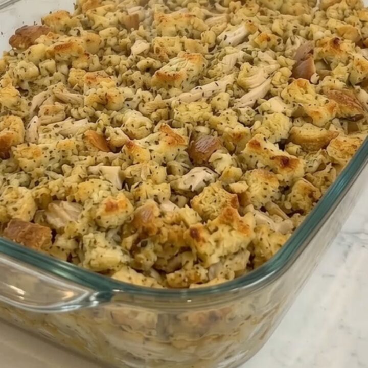 A golden-brown creamy chicken and herb stuffing bake in a glass dish on a marble countertop.