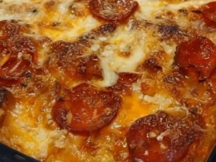 A close-up view of a serving spatula lifting a portion of hot, cheesy pizza casserole from a glass baking dish.
