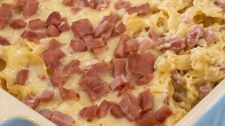 A warm, comforting Swiss ham and noodle casserole in a fluted blue dish, topped with melted golden cheese and diced pink ham.