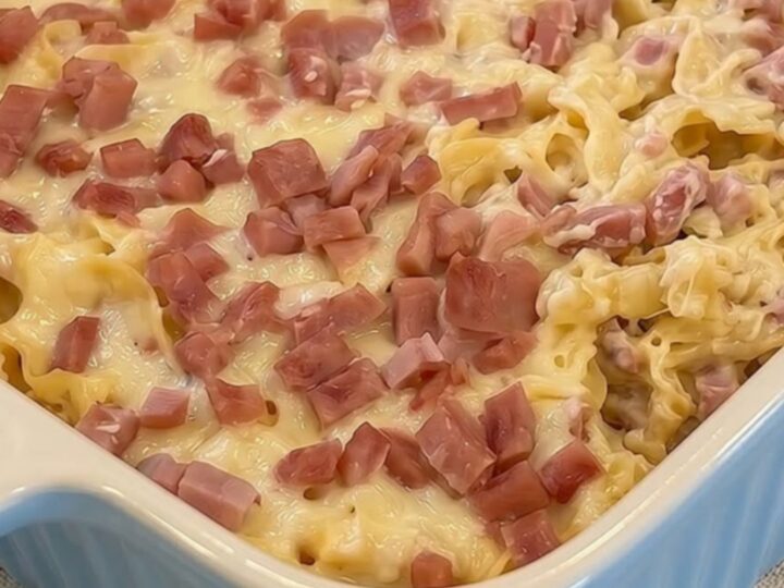A warm, comforting Swiss ham and noodle casserole in a fluted blue dish, topped with melted golden cheese and diced pink ham.