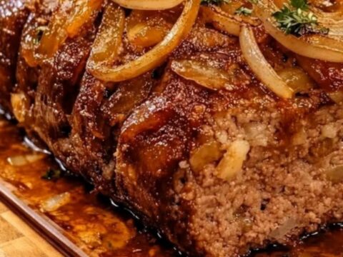 A juicy, tender slice of French onion meatloaf topped with rich beef glaze and golden caramelized onions on a wooden board.