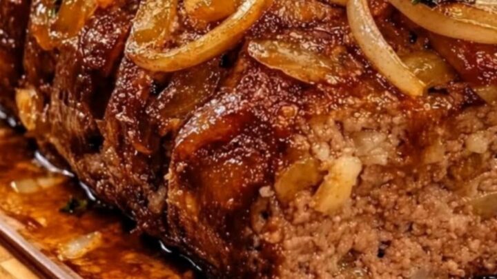 A juicy, tender slice of French onion meatloaf topped with rich beef glaze and golden caramelized onions on a wooden board.