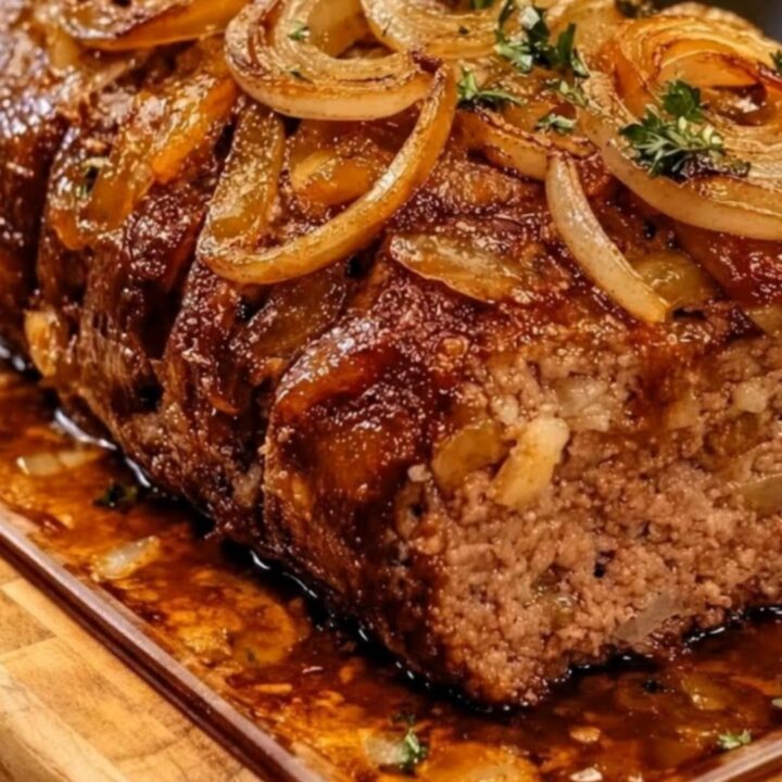 A juicy, tender slice of French onion meatloaf topped with rich beef glaze and golden caramelized onions on a wooden board.