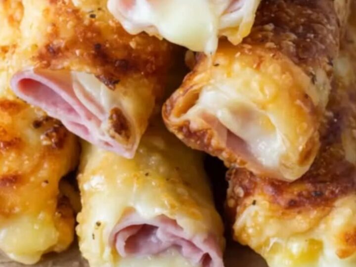 A rustic wooden board stacked with golden-brown baked ham and mozzarella rolls, showing a stretchy cheese pull.