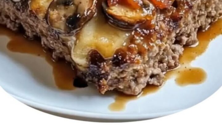 A rectangular meatloaf topped with melted Swiss cheese, sautéed mushrooms, bacon, and fresh thyme on a white plate.