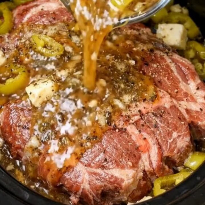 A glossy slow cooker pepperoncini pot roast shredded and surrounded by tangy peppers in a black slow cooker
