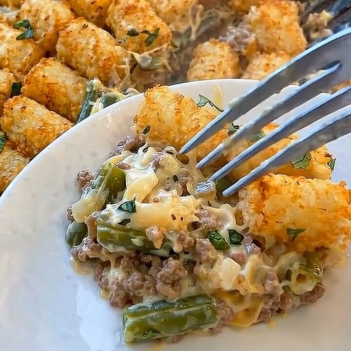 A golden, bubbling green bean tater tot casserole in a glass baking dish with a plated portion in the foreground.