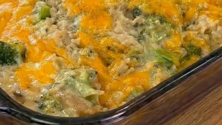 A steaming scoop of cheesy chicken broccoli rice casserole being lifted from a glass baking dish with a long cheese pull.