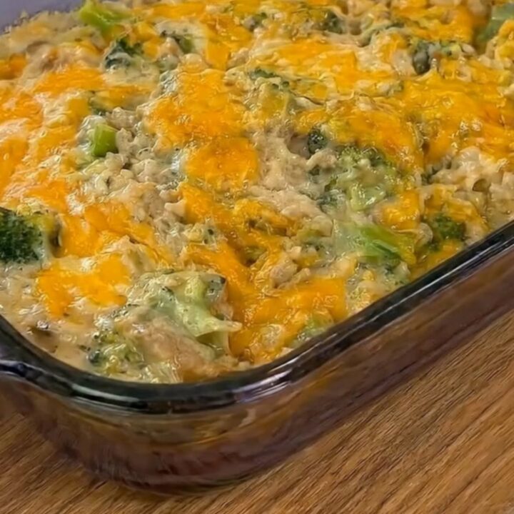 A steaming scoop of cheesy chicken broccoli rice casserole being lifted from a glass baking dish with a long cheese pull.