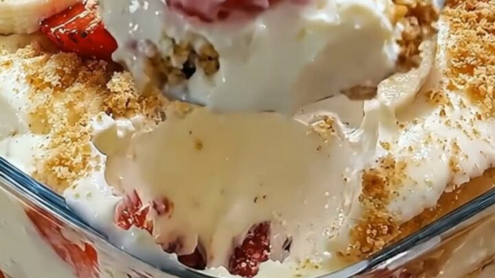 A close-up of a glass filled with layers of vanilla pudding, sliced strawberries, bananas, and a golden cookie crumb topping.