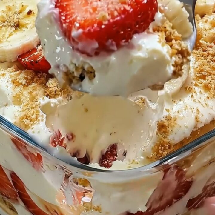 A close-up of a glass filled with layers of vanilla pudding, sliced strawberries, bananas, and a golden cookie crumb topping.