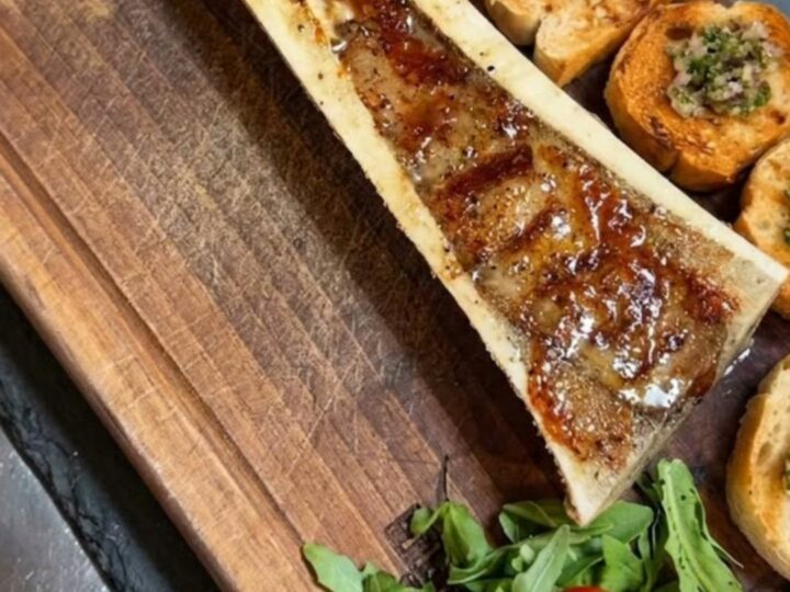 Vue en gros plan d'une moelle de bœuf rôtie servie sur une planche en bois avec des crostini et de la roquette.