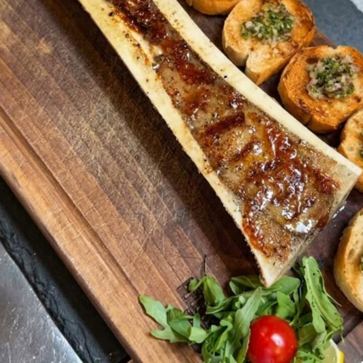 Vue en gros plan d'une moelle de bœuf rôtie servie sur une planche en bois avec des crostini et de la roquette.