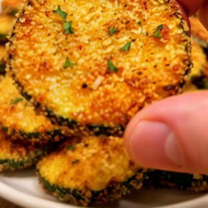 A pile of golden, crispy parmesan zucchini chips dusted with fresh thyme leaves on a white plate.