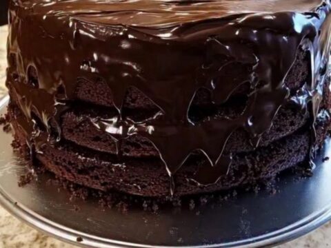 A close-up of a three-layer chocolate cake on a silver pedestal stand with a glossy ganache finish.