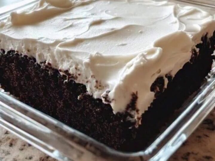 A slice of moist chocolate cake in a square glass dish with fluffy white frosting being swirled on top.