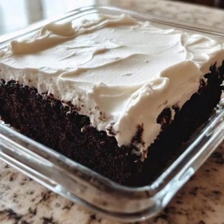A slice of moist chocolate cake in a square glass dish with fluffy white frosting being swirled on top.