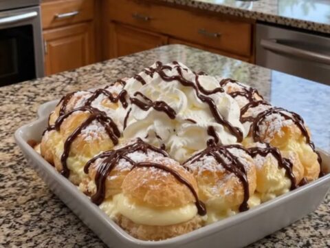A gorgeous layered cream puff cake topped with whipped cream, powdered sugar, and a glossy chocolate drizzle.