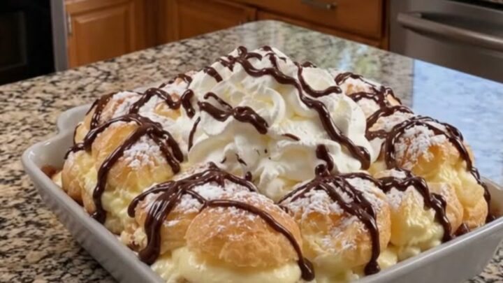 A gorgeous layered cream puff cake topped with whipped cream, powdered sugar, and a glossy chocolate drizzle.