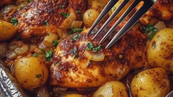 A rustic wooden table holding a glistening aluminum foil pan filled with roasted garlic butter chicken and golden baby