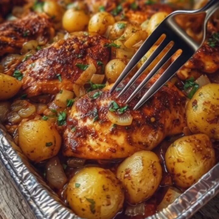A rustic wooden table holding a glistening aluminum foil pan filled with roasted garlic butter chicken and golden baby
