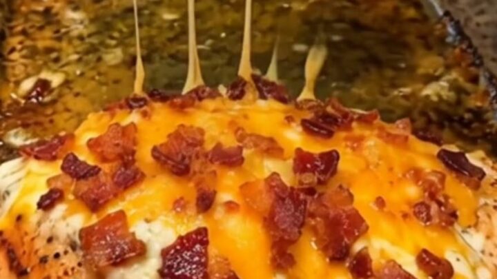 A baked chicken breast being lifted from a clear glass baking dish, showing a long melt of cheddar cheese and bacon bits.