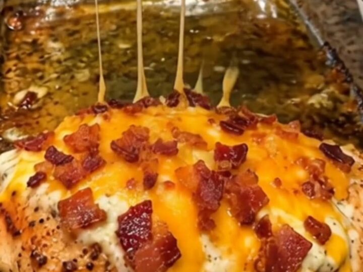 A baked chicken breast being lifted from a clear glass baking dish, showing a long melt of cheddar cheese and bacon bits.