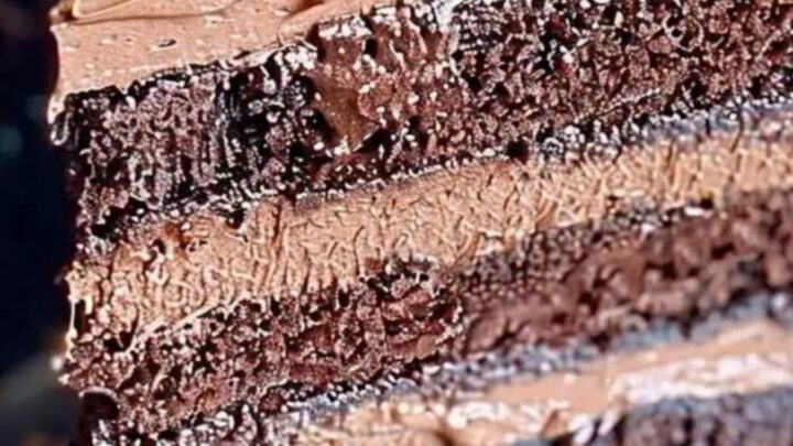 A slice of triple-layer chocolate cake with whipped frosting and shavings on a dark plate.