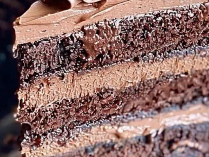 A slice of triple-layer chocolate cake with whipped frosting and shavings on a dark plate.