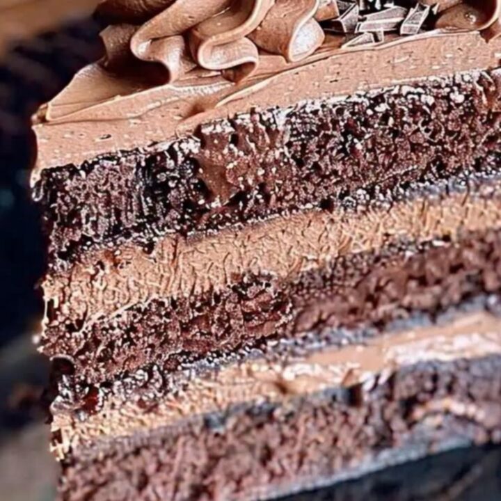 A slice of triple-layer chocolate cake with whipped frosting and shavings on a dark plate.