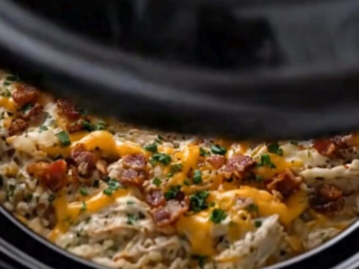 A serving spoon lifting creamy shredded crack chicken with melted cheddar and bacon from a slow cooker.