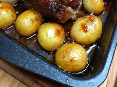 A golden, honey-glazed pork roast served in a black pan with roasted baby potatoes and caramelized onions.