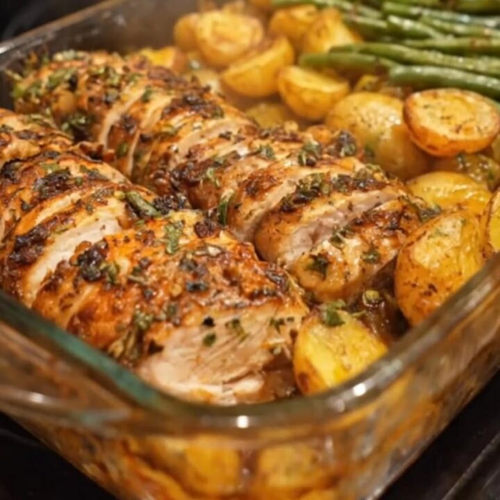 A golden roasted chicken breast sliced next to roasted baby potatoes and tender green beans in a glass dish.