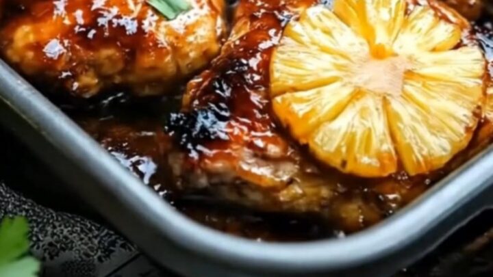 A close-up of sticky pineapple chicken with caramelized fruit in a roasting pan with steam rising.