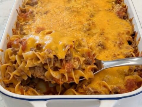 A close-up of a metal spoon lifting a scoop of cheesy beef egg noodles with melting cheddar cheese stretching from the white