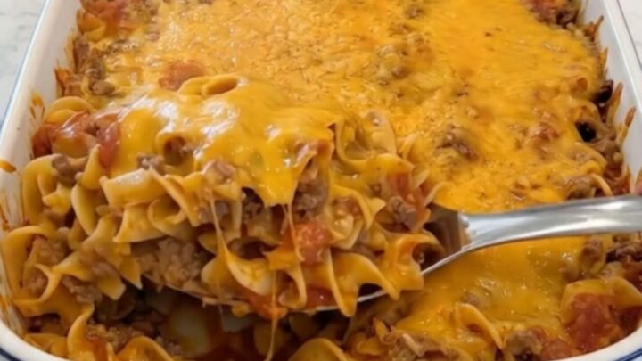 A close-up of a metal spoon lifting a scoop of cheesy beef egg noodles with melting cheddar cheese stretching from the white