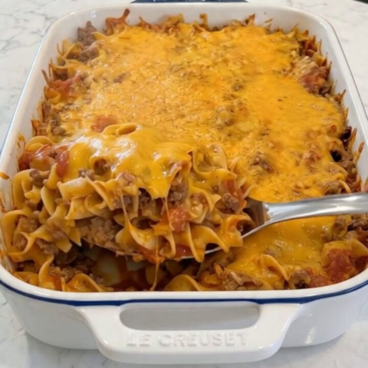 A close-up of a metal spoon lifting a scoop of cheesy beef egg noodles with melting cheddar cheese stretching from the white