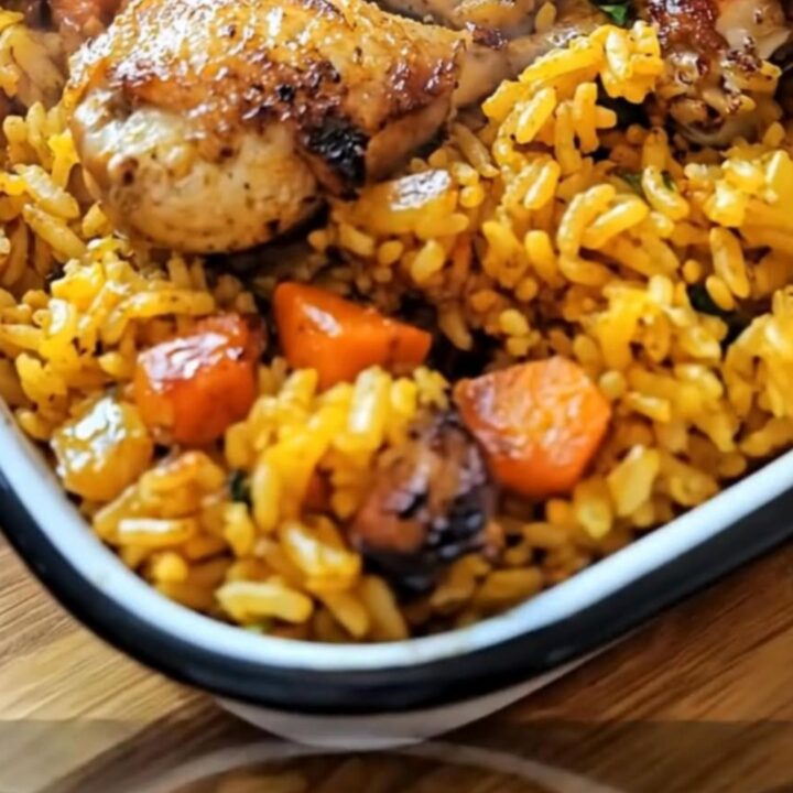 A golden roasted chicken thigh resting on a bed of fluffy yellow rice with carrots in a white ceramic dish.