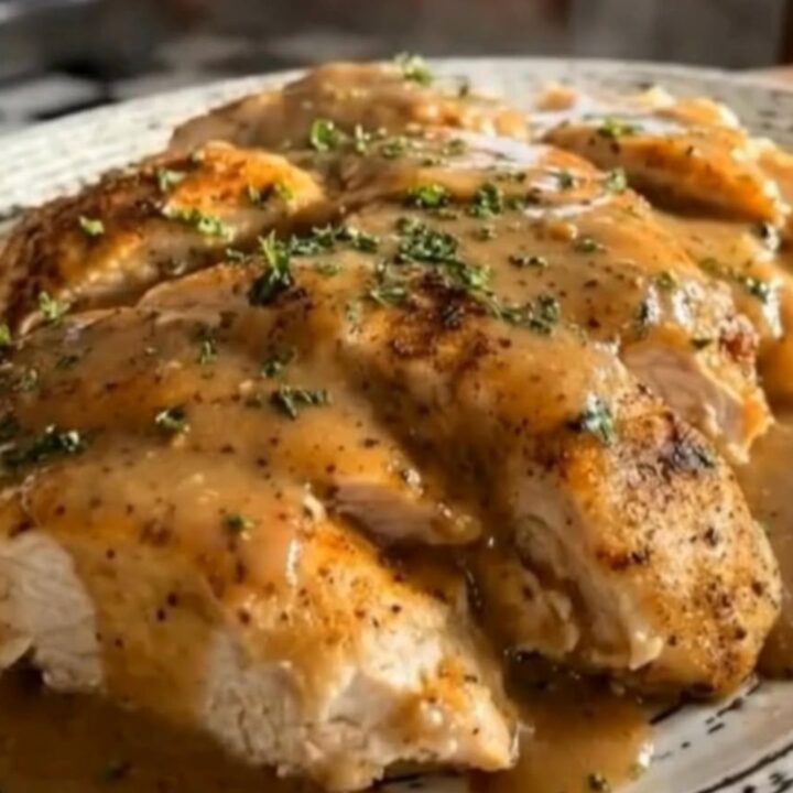 Tender chicken breasts covered in savory brown gravy on a ceramic platter, garnished with fresh parsley.
